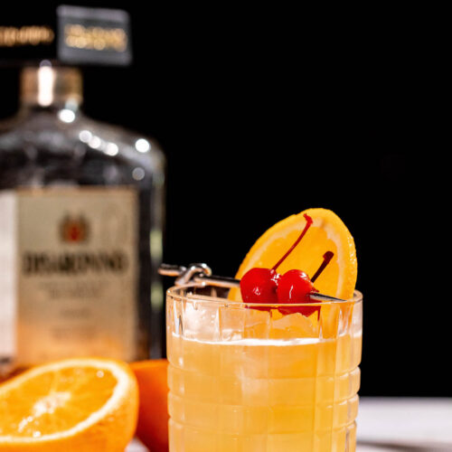 An amaretto stone sour cocktail garnished with an orange slice and cherries sits in a glass, with a bottle and sliced orange in the background.