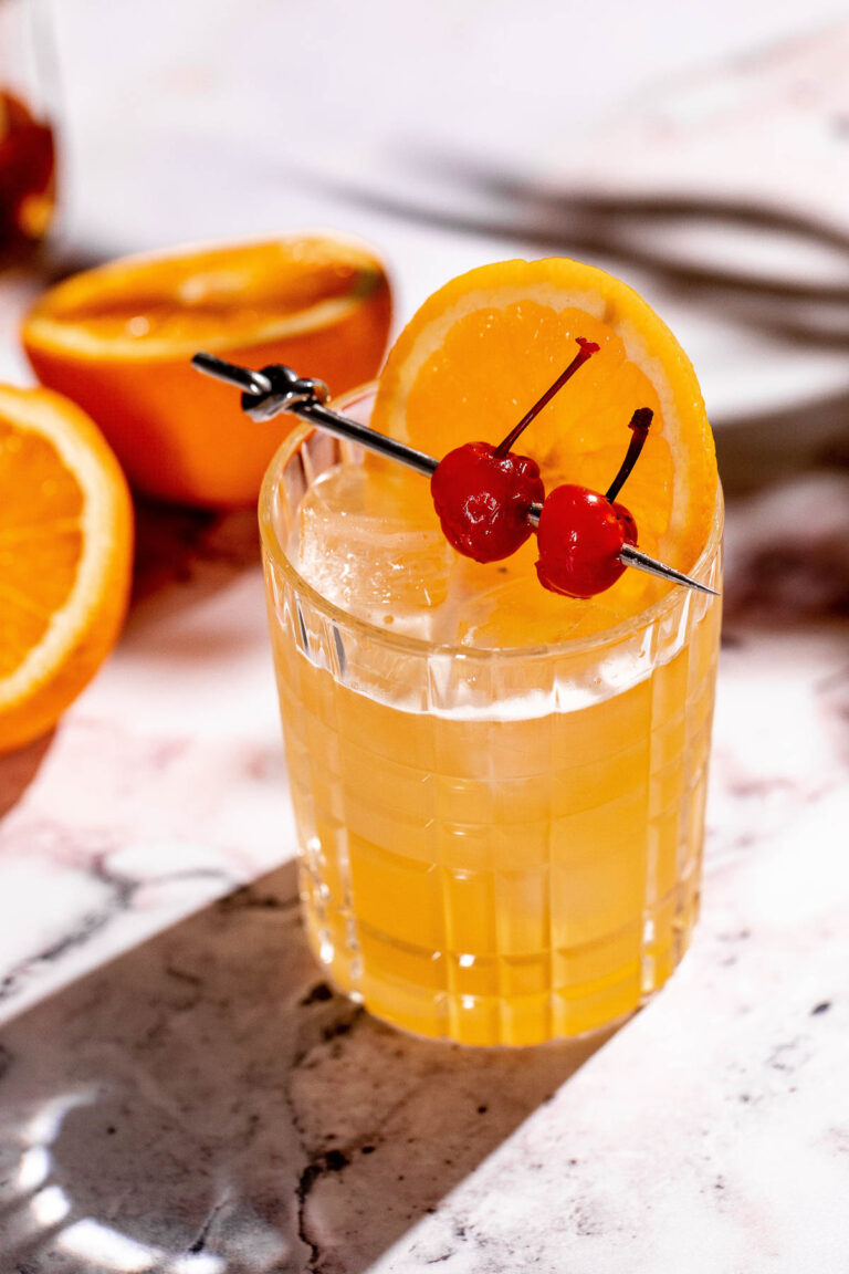 A glass of iced amaretto stone sour drink garnished with an orange slice and two cherries on a skewer sits on a marble surface with sliced oranges nearby.