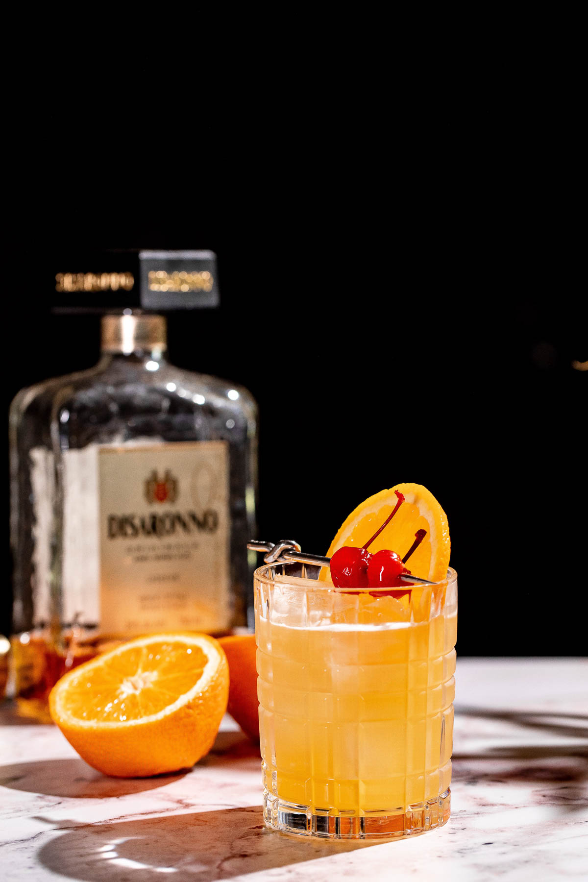 A glass of amaretto stone sour with an orange slice and cherries sits on a marble surface, with a bottle of Disaronno and sliced oranges in the background.