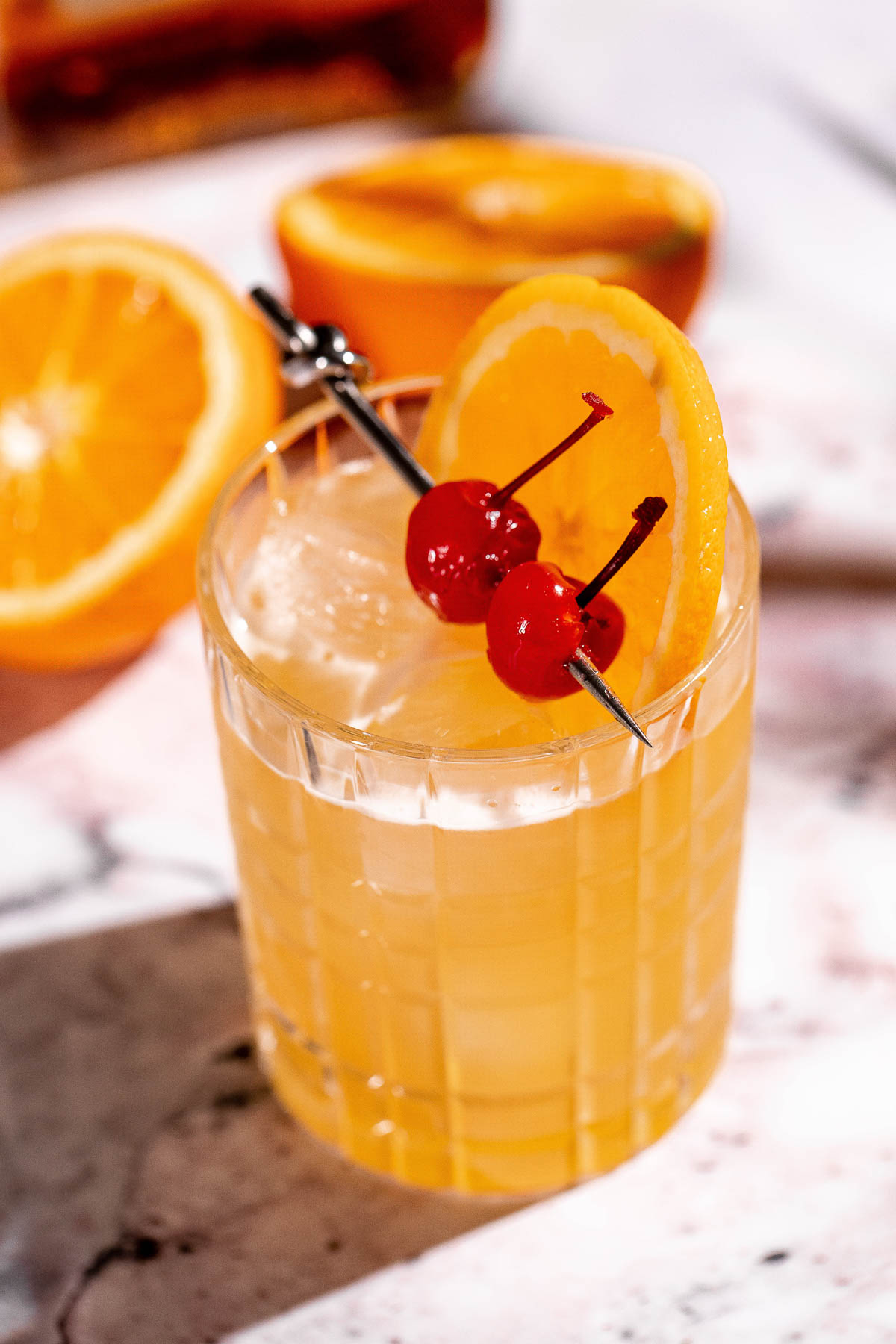 An amaretto stone sour cocktail served in a clear glass with ice cubes, garnished with two cherries and an orange slice, is set against halved oranges in the background.