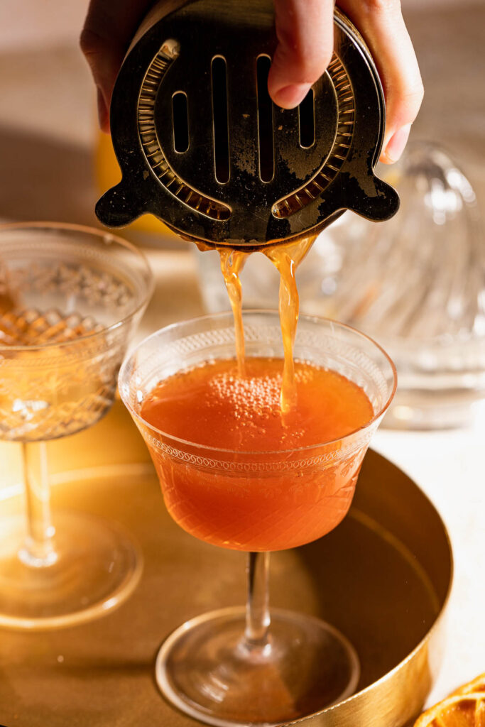 A hand pours an orange Blood and Sand cocktail through a strainer into a glass on a tray, with another glass in the background.