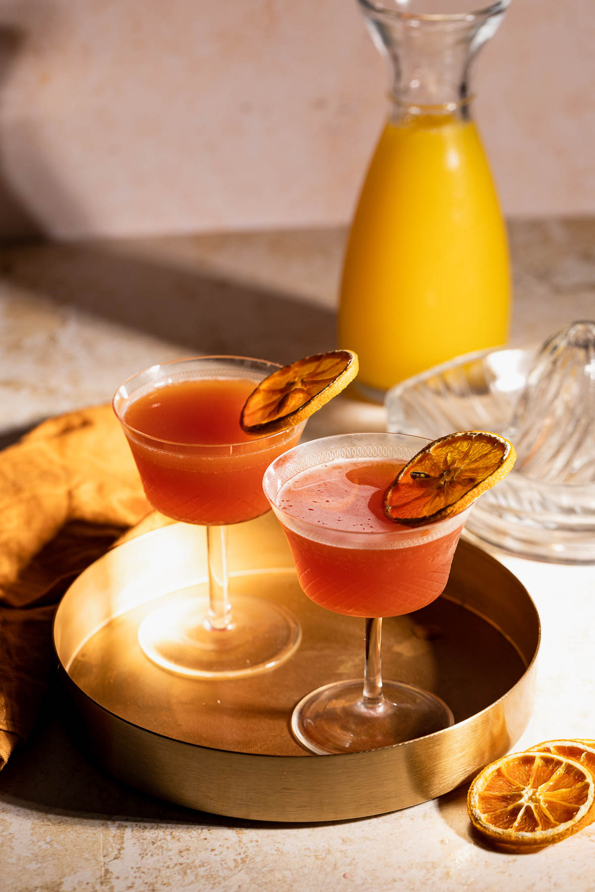 Two cocktail glasses with pink Blood and Sand cocktails and dried orange garnish sit on a golden tray, with a carafe of orange juice and a citrus juicer in the background.