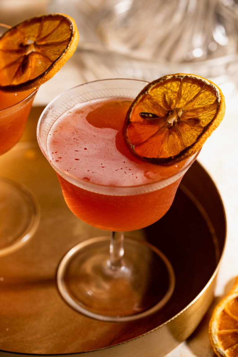 A pink Blood and Sand cocktail with a dried orange slice garnish rests on a brass tray.