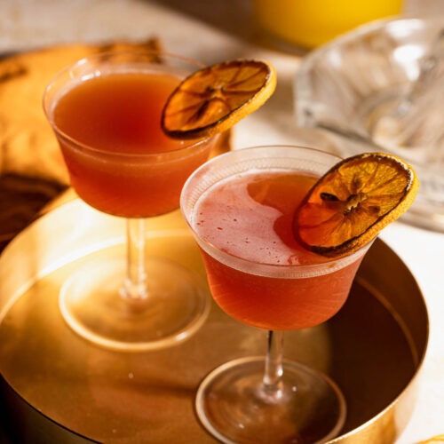 Two cocktail glasses filled with a red-orange Blood and Sand drink and garnished with dried orange slices sit on a round, gold tray.