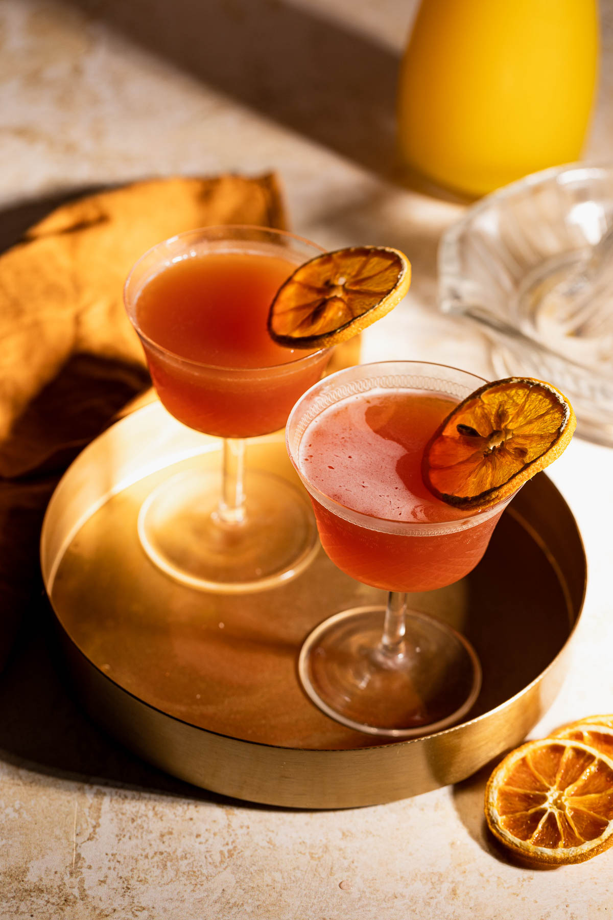 Two cocktail glasses with pink Blood and Sand drinks and dried orange garnishes stand on a round golden tray, with a bottle and cloth in the background.