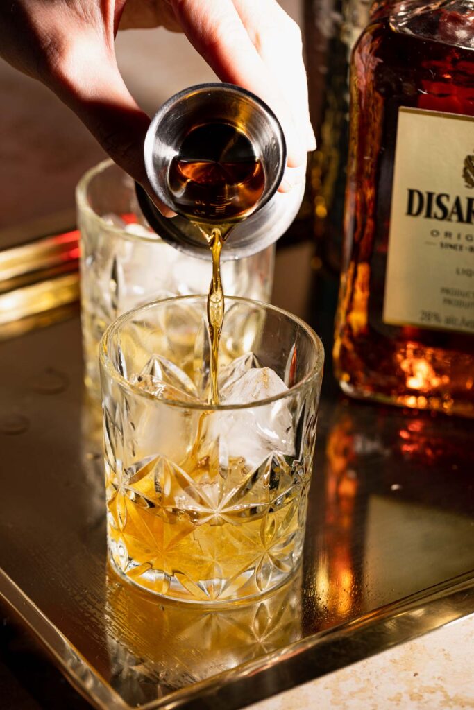 A hand pours amaretto from a jigger into a glass on a tray beside a Disaronno bottle.