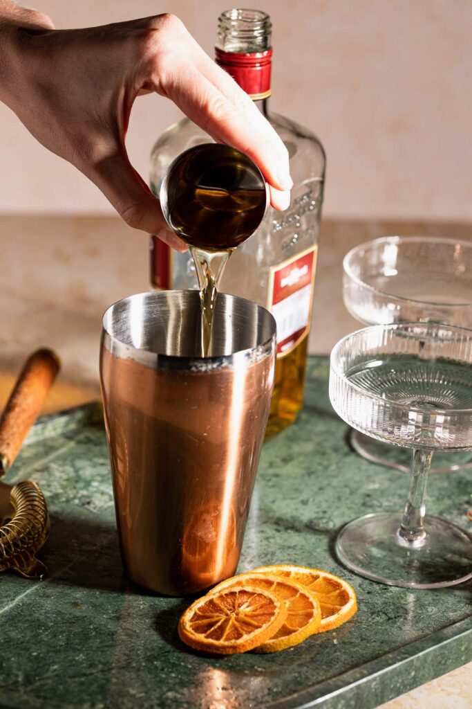 A hand pours bourbon from a small cup into a copper cocktail shaker beside two empty glasses and dried orange slices.