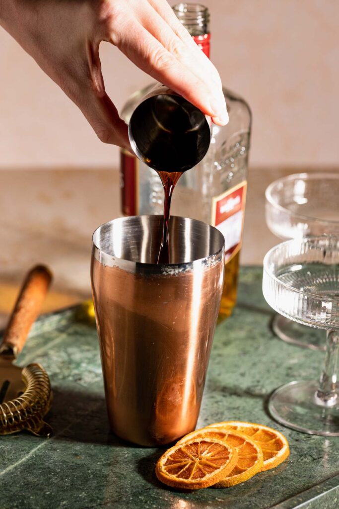 A hand pours coffee liquor from a jigger into a copper cocktail shaker on green marble, with dried orange slices and cocktail glasses in the background.