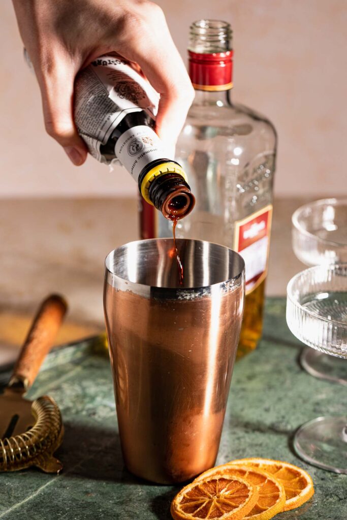 A hand pours bitters into a copper cocktail shaker surrounded by a bottle, cocktail glasses, and orange slices.