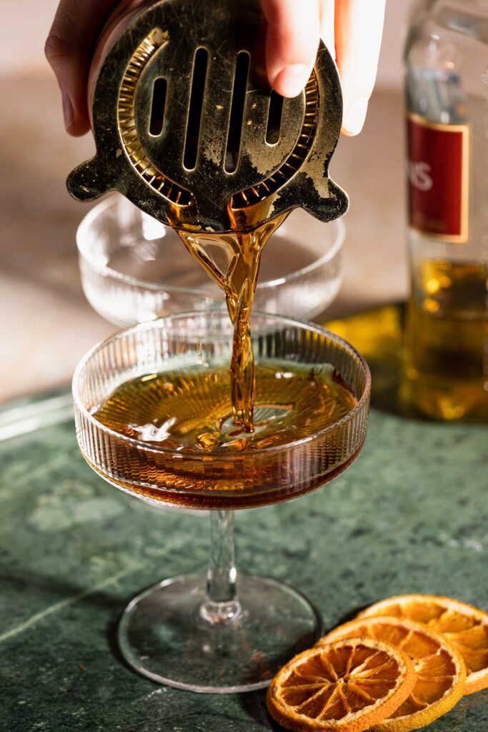 A hand pours the revolver cocktail through a strainer into a glass with dried citrus slices on the table and a partially visible bottle in the background.