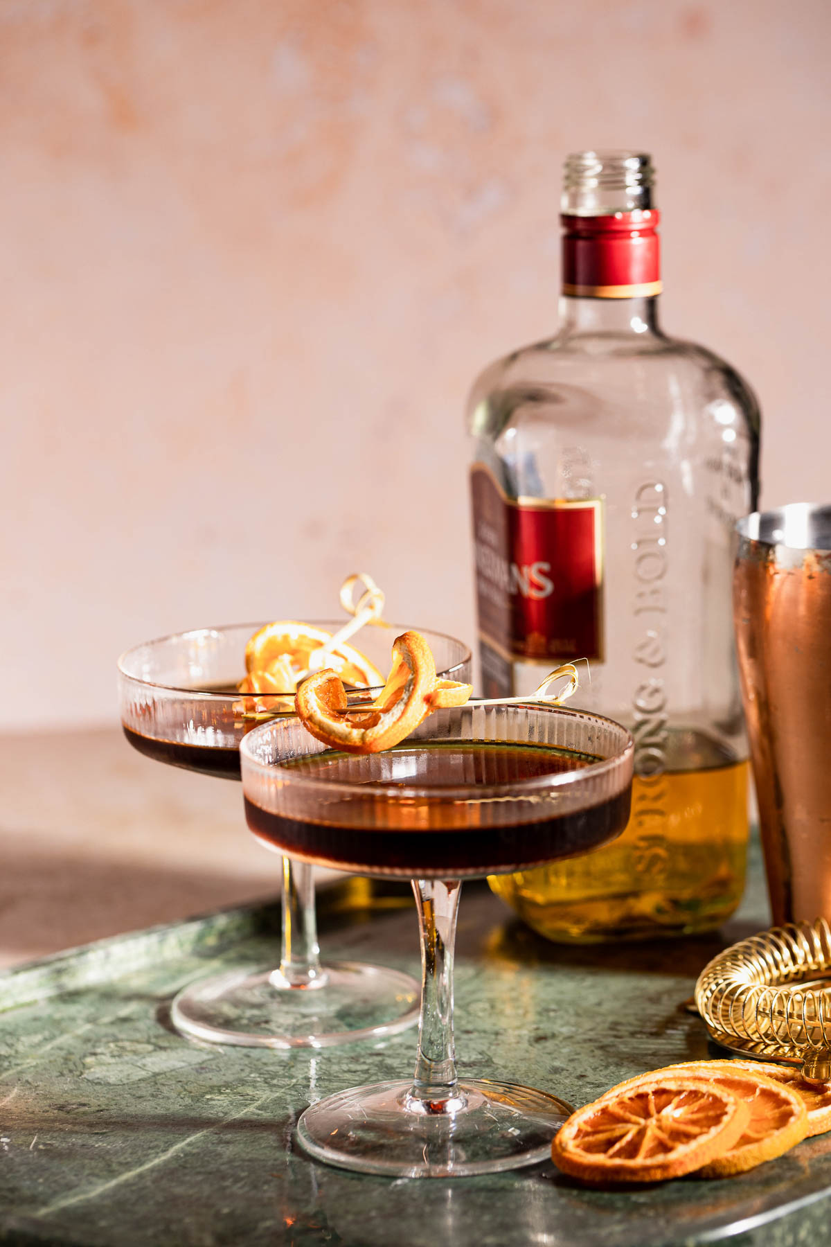 Two coupe glasses with a dark revolver cocktail sit on a green marble surface, accompanied by a bottle and dried citrus slices.