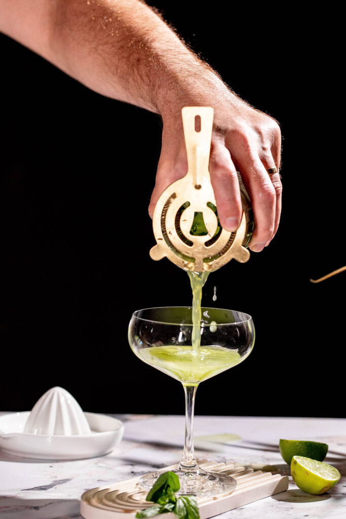 A hand strains a basil martini cocktail into a coupe glass on a marble counter with limes and a citrus squeezer.