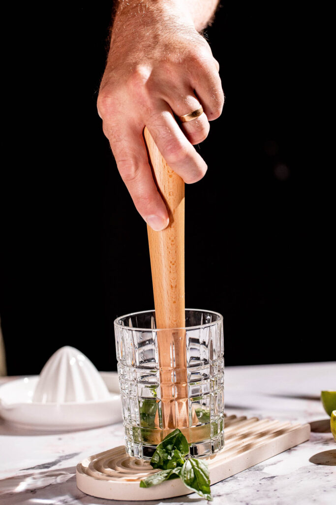 A hand muddles a glass of basil leaves on marble, surrounded by basil leaves and a citrus juicer.