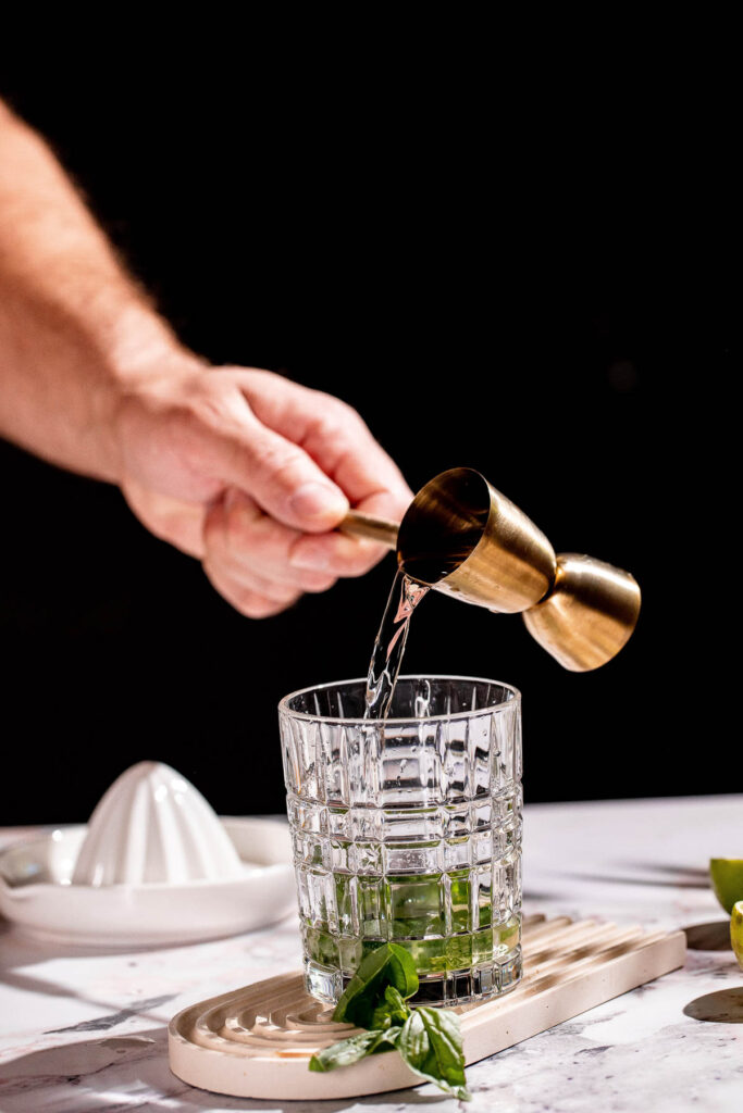 A person pours vodka from a brass jigger into a glass on a marble surface beside basil leaves.