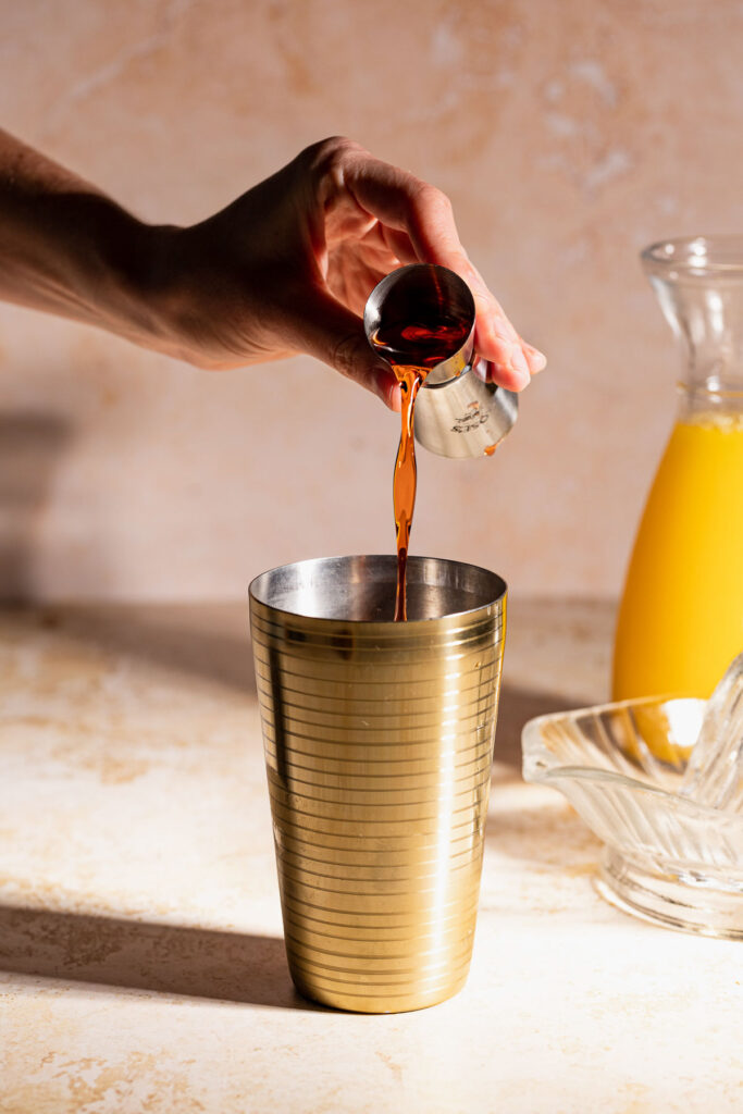 A person pours cherry liqueu from a measuring cup into a gold cocktail shaker on a countertop, with a glass pitcher of orange juice in the background.