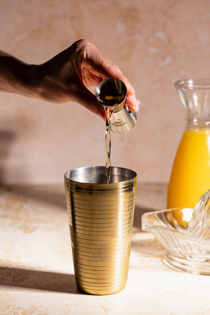 A hand pours sweet vermouth from a jigger into a gold cocktail shaker on a countertop beside a glass orange juice pitcher and juicer.