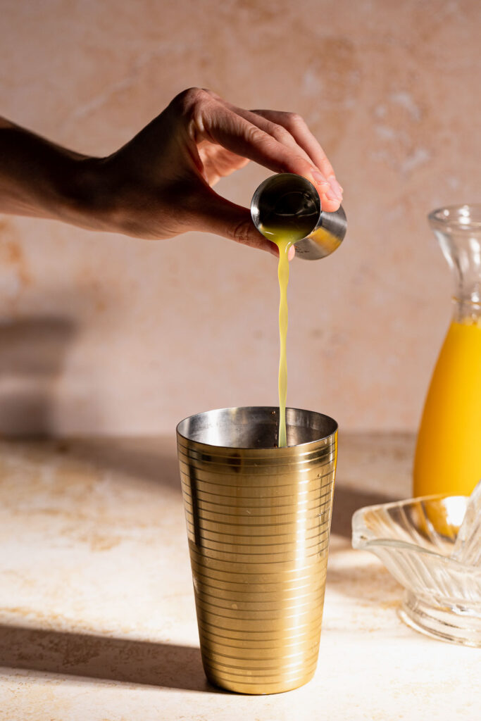 A hand pours orange juice from a jigger into a gold cocktail shaker, with an orange juice jug and glass plate in the background.