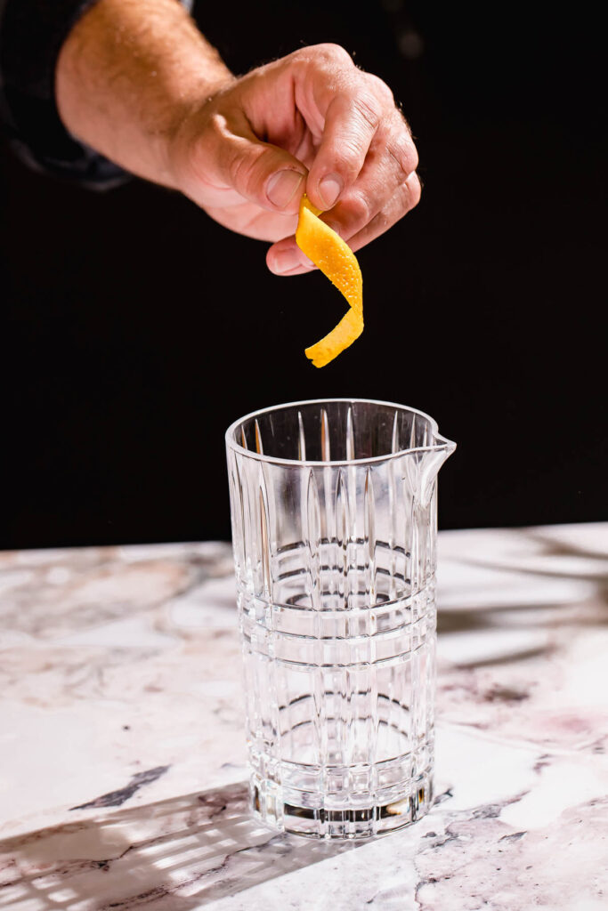 A hand holds a citrus peel above an empty faceted glass on marble.