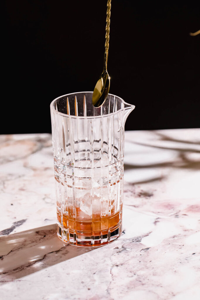 A glass of brown liquid with ice on a marble surface has a metal spoon hovering above it.