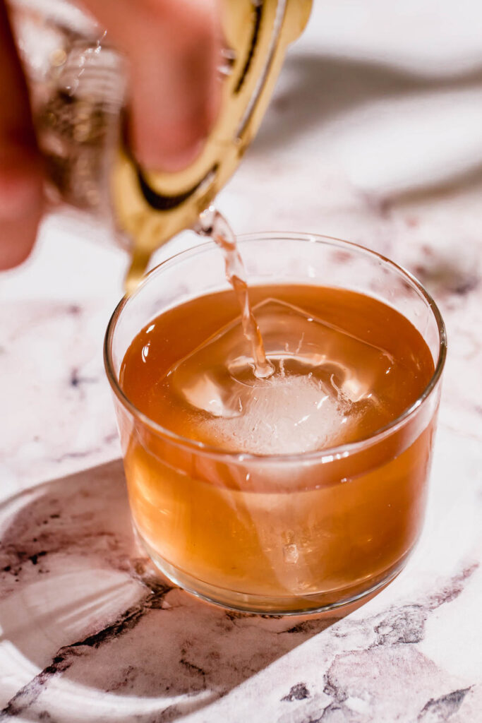 A hand pours a tequila old fashioned from a shaker into a glass with a large ice cube on marble.