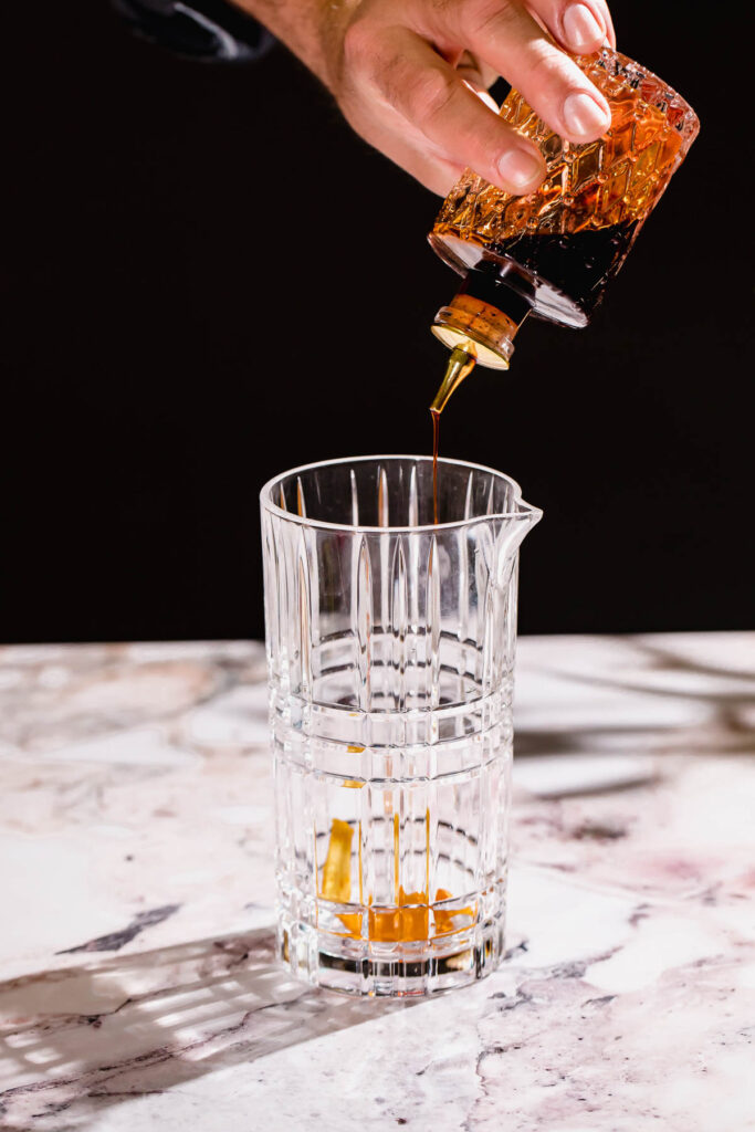 A hand pours bitters from a decorative glass container into a tall pitcher on a marble surface.