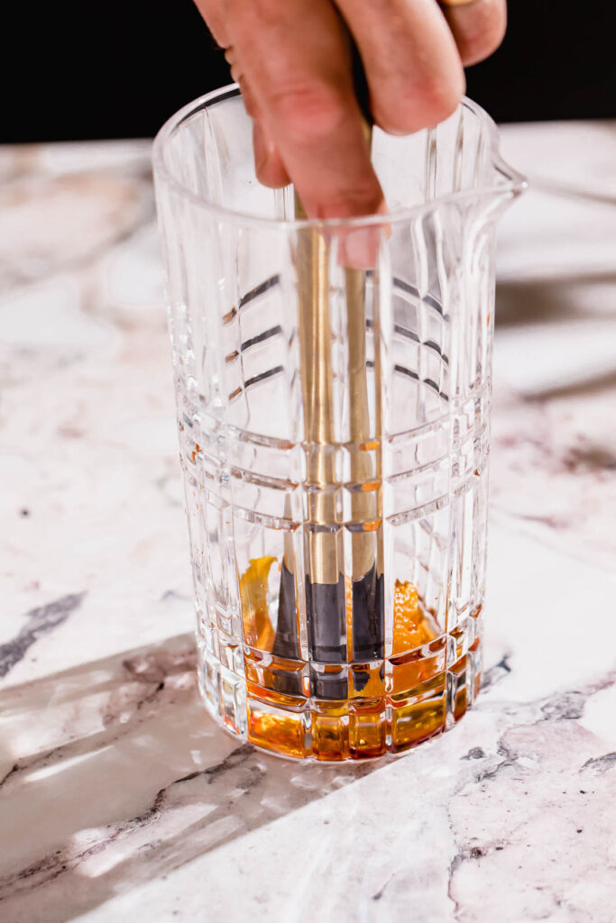 Using a muddler, someone stirs a drink with orange slices in a textured glass on a marble countertop.