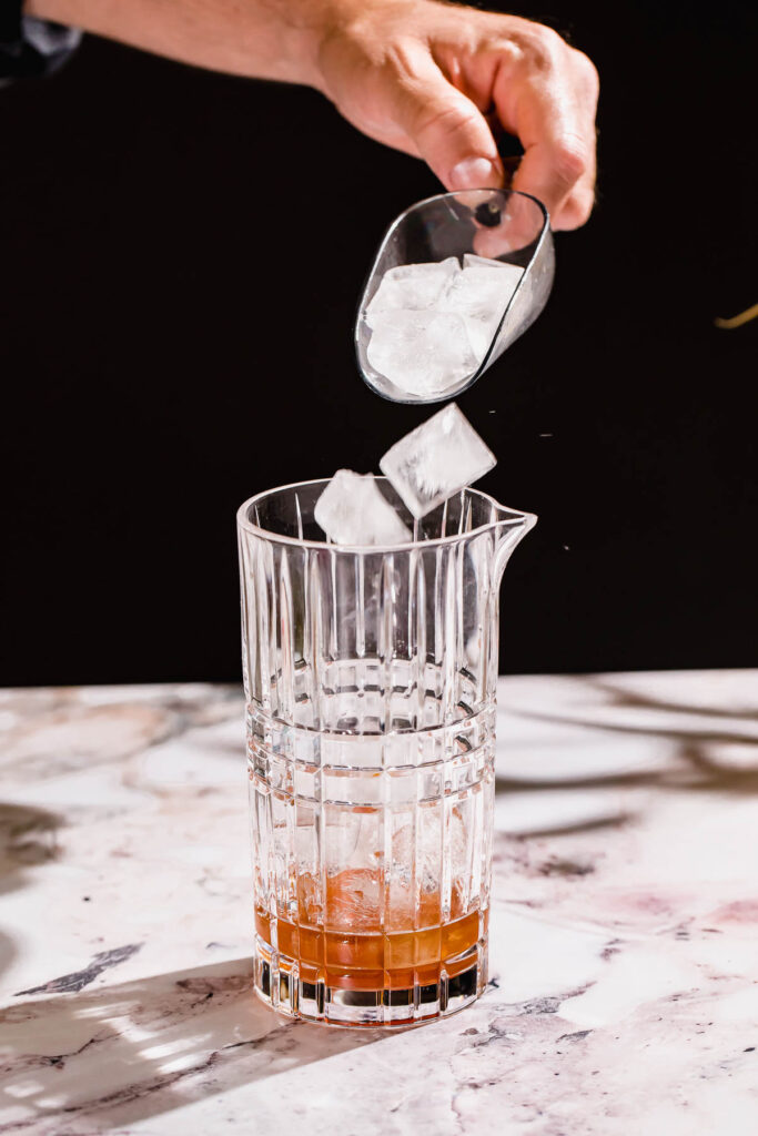 A hand adds ice cubes to a partly filled glass mixing container on a marble surface.