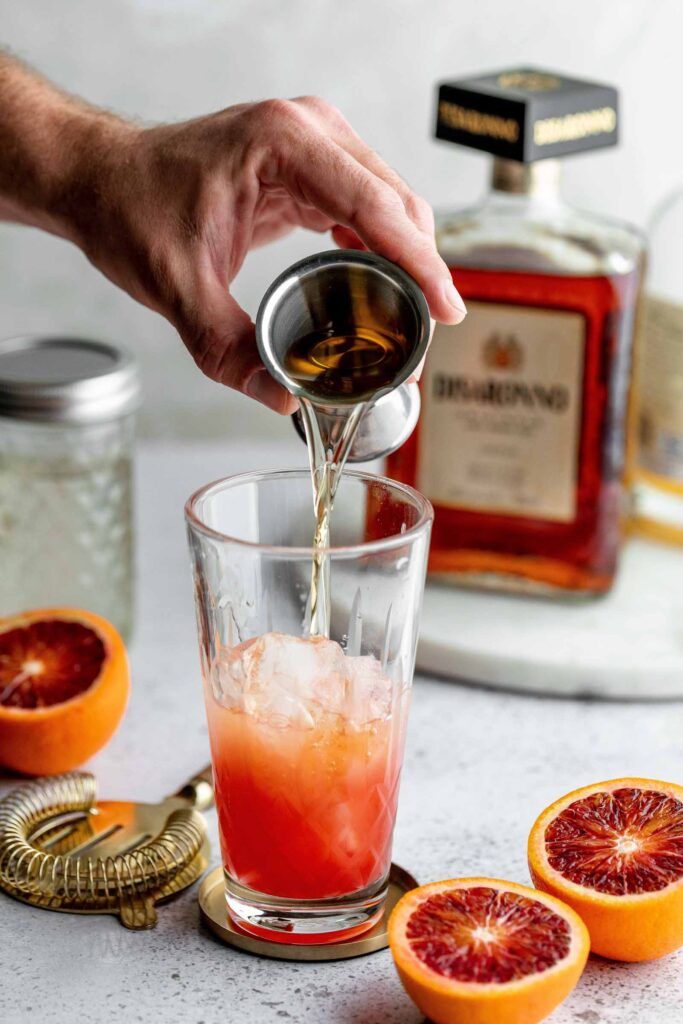 A hand pours liquid from a metal jigger into an ice-filled glass with red-orange juice, set against blood oranges and a bottle in the background.