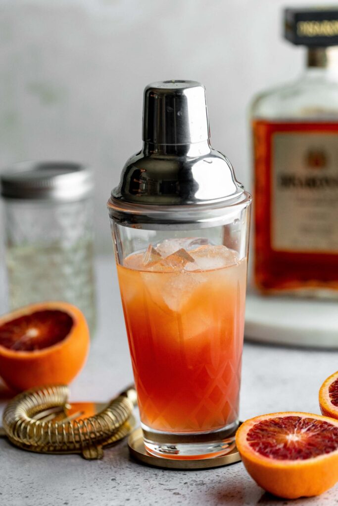 A cocktail shaker with an orange-red drink and ice sits amid halved blood oranges, a strainer, and a bottle on a gray countertop.