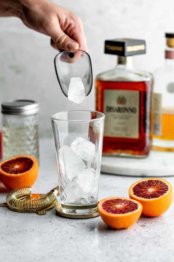 A hand places ice cubes into a glass on a counter with blood oranges, a strainer, and bottles of liquor.