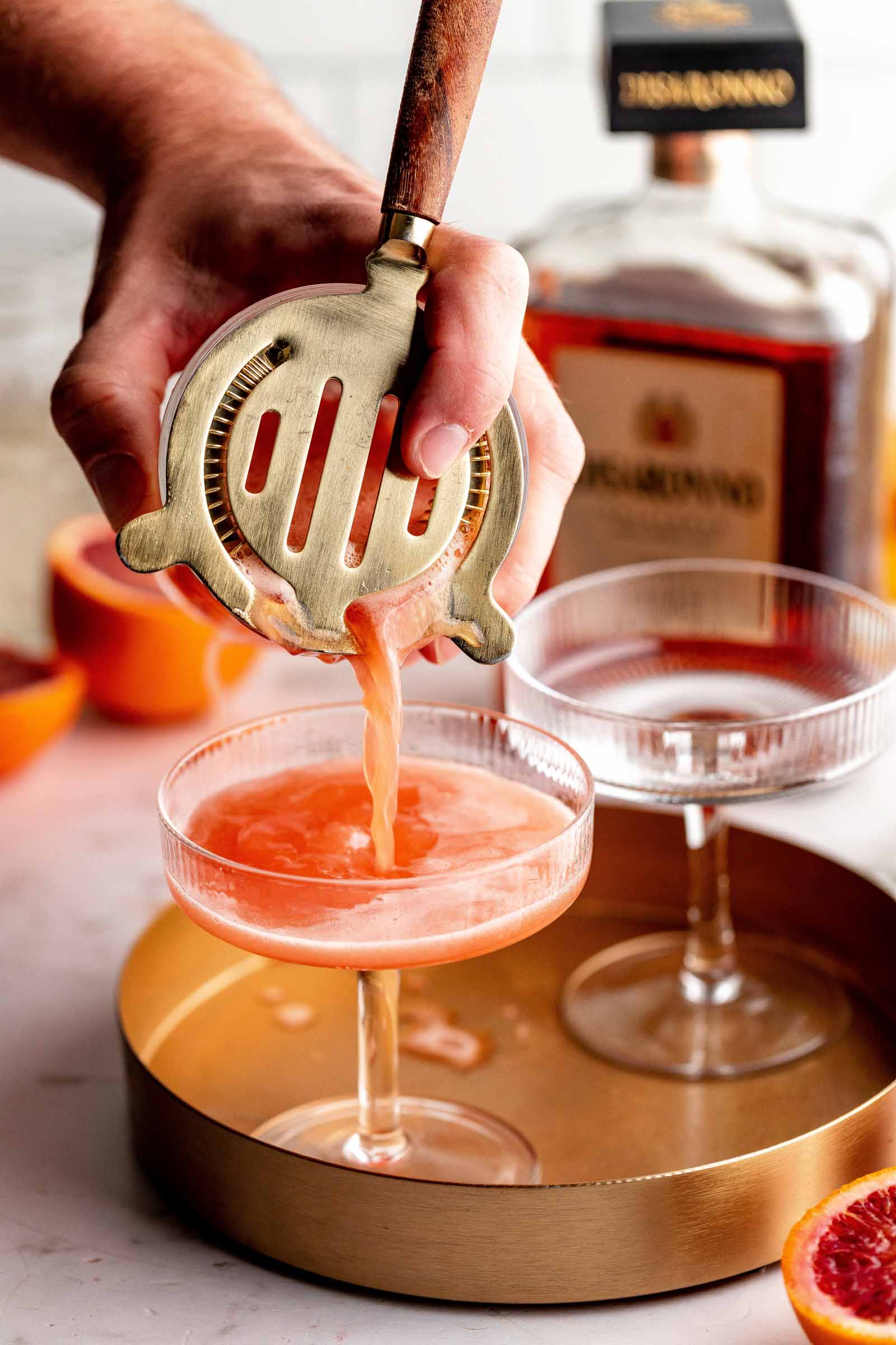 A hand pours an amaretto sour cocktail through a strainer into a glass on a tray, with a bottle and sliced grapefruit in the background.