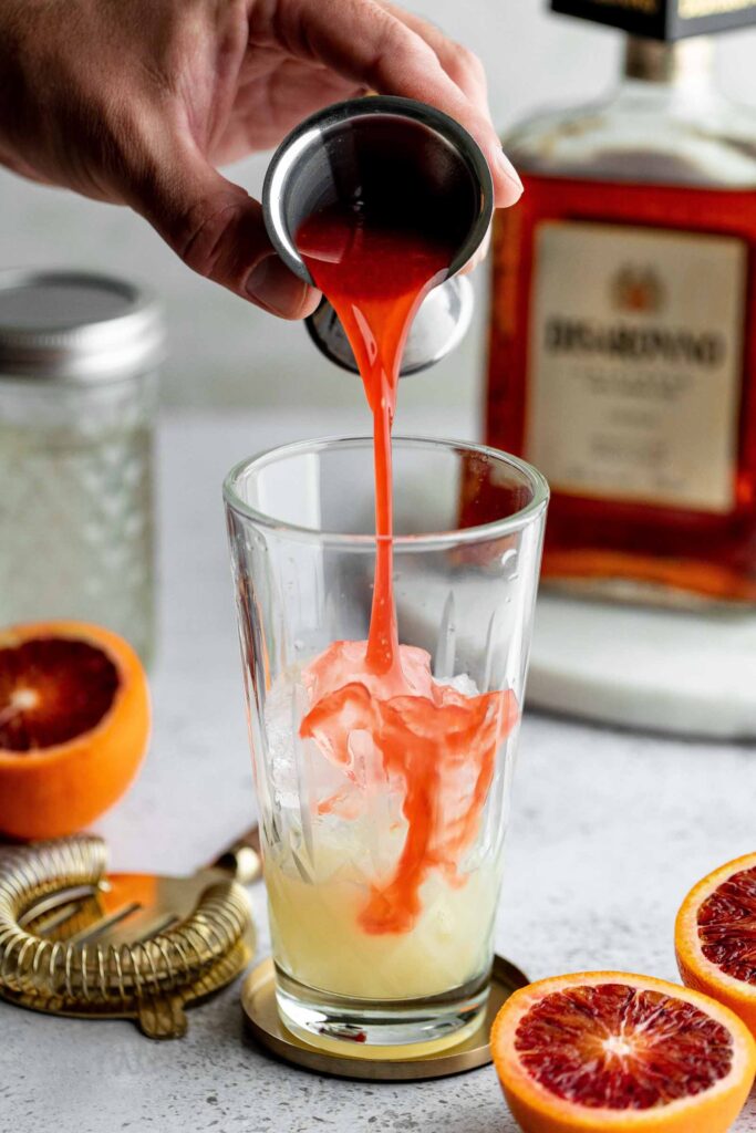 Blood orange juice is poured into a glass with ice, with sliced oranges and a bottle in the background.