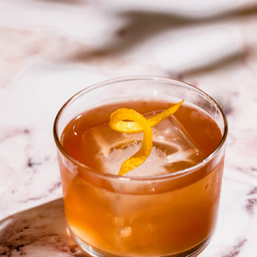 A tequila old fashioned cocktail with a large ice cube and lemon peel sits on marble.