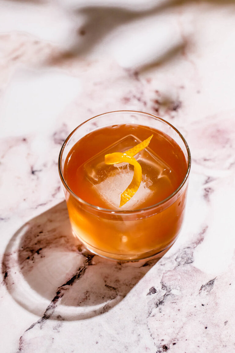 A tequila old fashioned cocktail with a lemon twist and a large ice cube sits on a marbled surface.