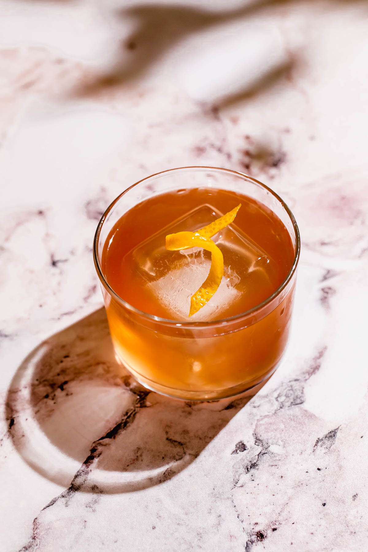 A tequila cocktail with ice cubes and a lemon twist garnish sits on a marble surface.