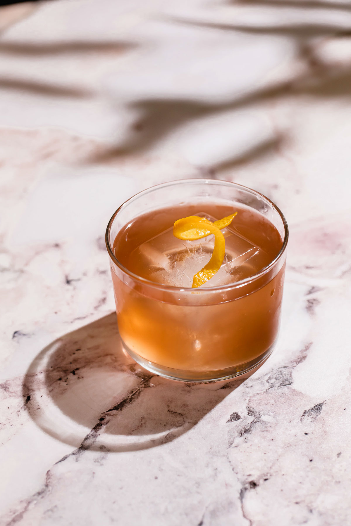 A tequila old fashioned cocktail with a large ice cube and lemon peel garnish rests on a marble surface.