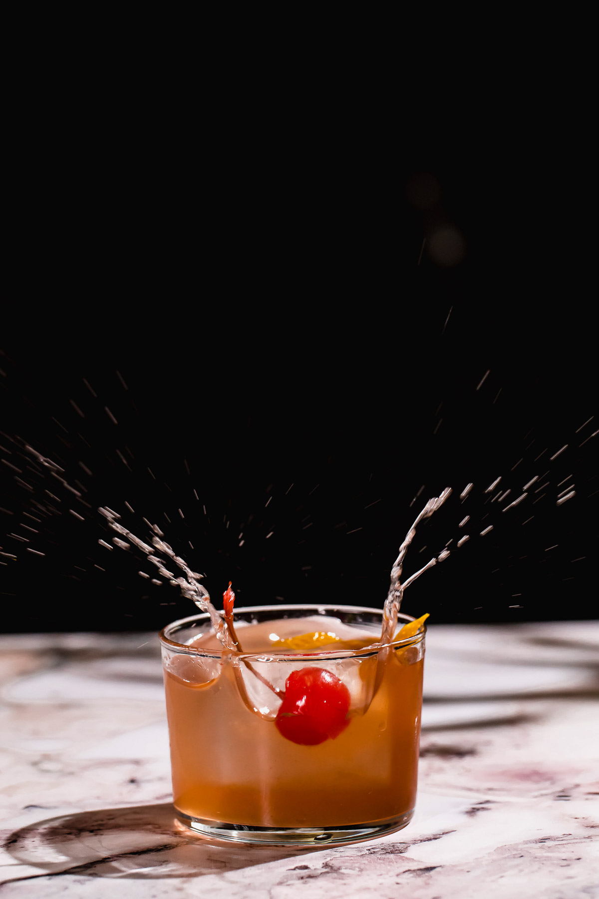 A splash occurs in a tequila old fashioned with ice, cherry, and orange against a dark background.