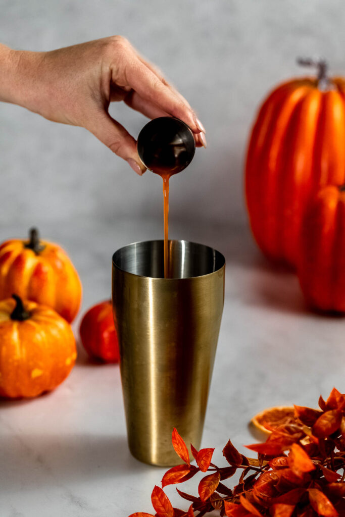 A hand pours pumpkin spice syrup from a small cup into a metal tumbler against a background of three pumpkins and orange leaves.