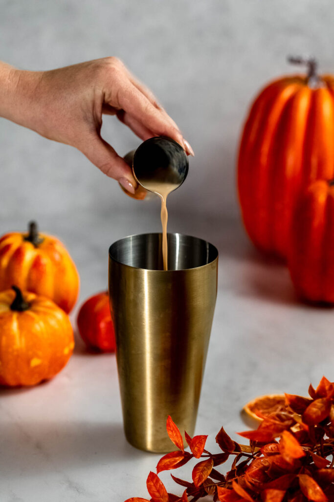 A hand pours irish cream from a small cup into a metal shaker with pumpkins and orange leaves in the background.