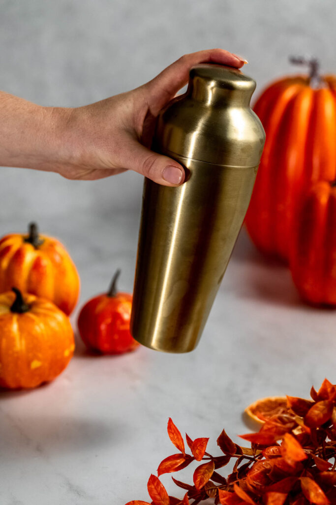 A hand holds a brass cocktail shaker in front of small pumpkins and autumn leaves on marble.