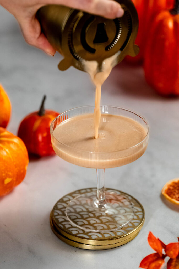 A hand pours a creamy baileys pumpkin spice martini cocktail from a strainer into a glass, surrounded by small pumpkins and orange slices on marble.