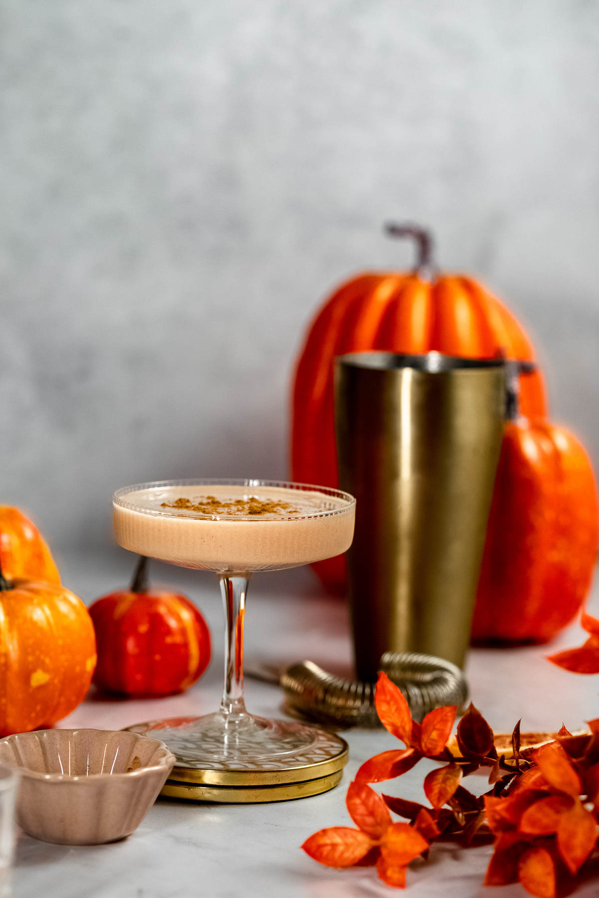 A coupe glass filled with a baileys pumpkin spice martini sits on a table surrounded by pumpkins, a shaker, and orange leaves.