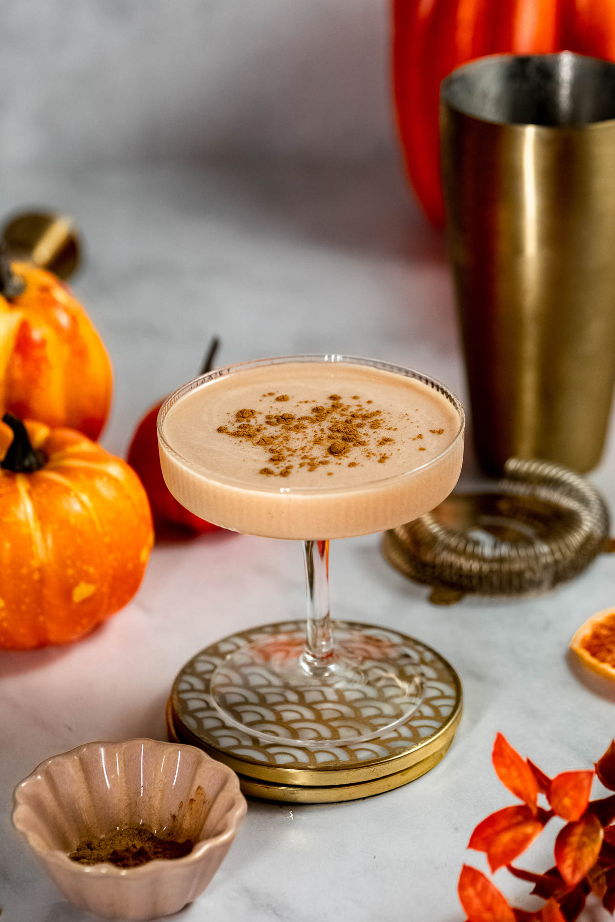 A creamy baileys pumpkin spice martini cocktail sits in a glass on a decorative coaster, surrounded by small pumpkins, an empty shaker, a strainer, and orange foliage on marble.