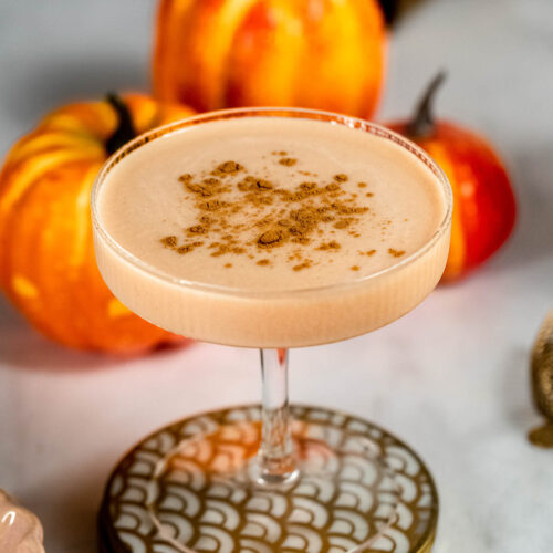 A cocktail glass filled with a creamy baileys pumpkin spice martini topped with spices is set against a background of decorative pumpkins.