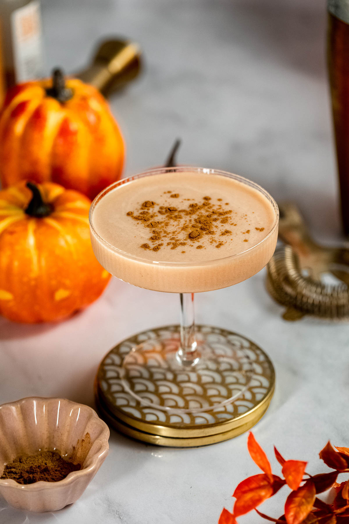 A creamy baileys pumpkin spice martini garnished with ground spice sits on a patterned coaster beside small pumpkins and a bowl of spice on a marble surface.