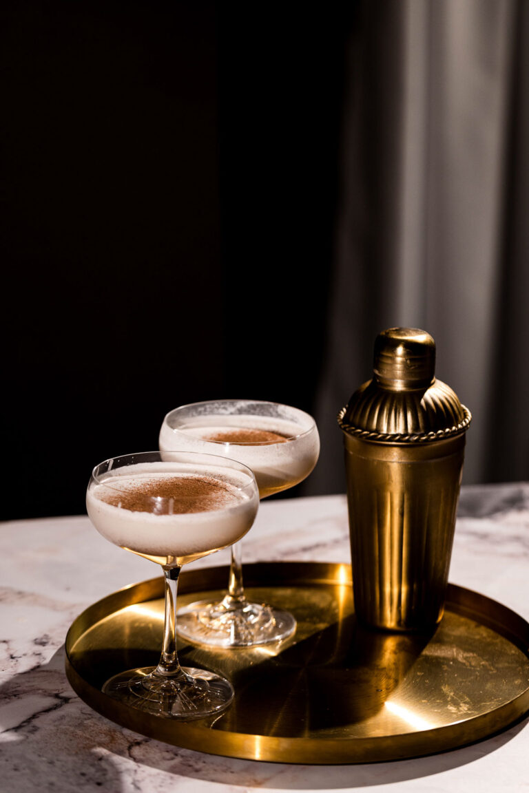 Two creamy eggnog martinis in glasses sit on a gold tray next to a shaker on marble.