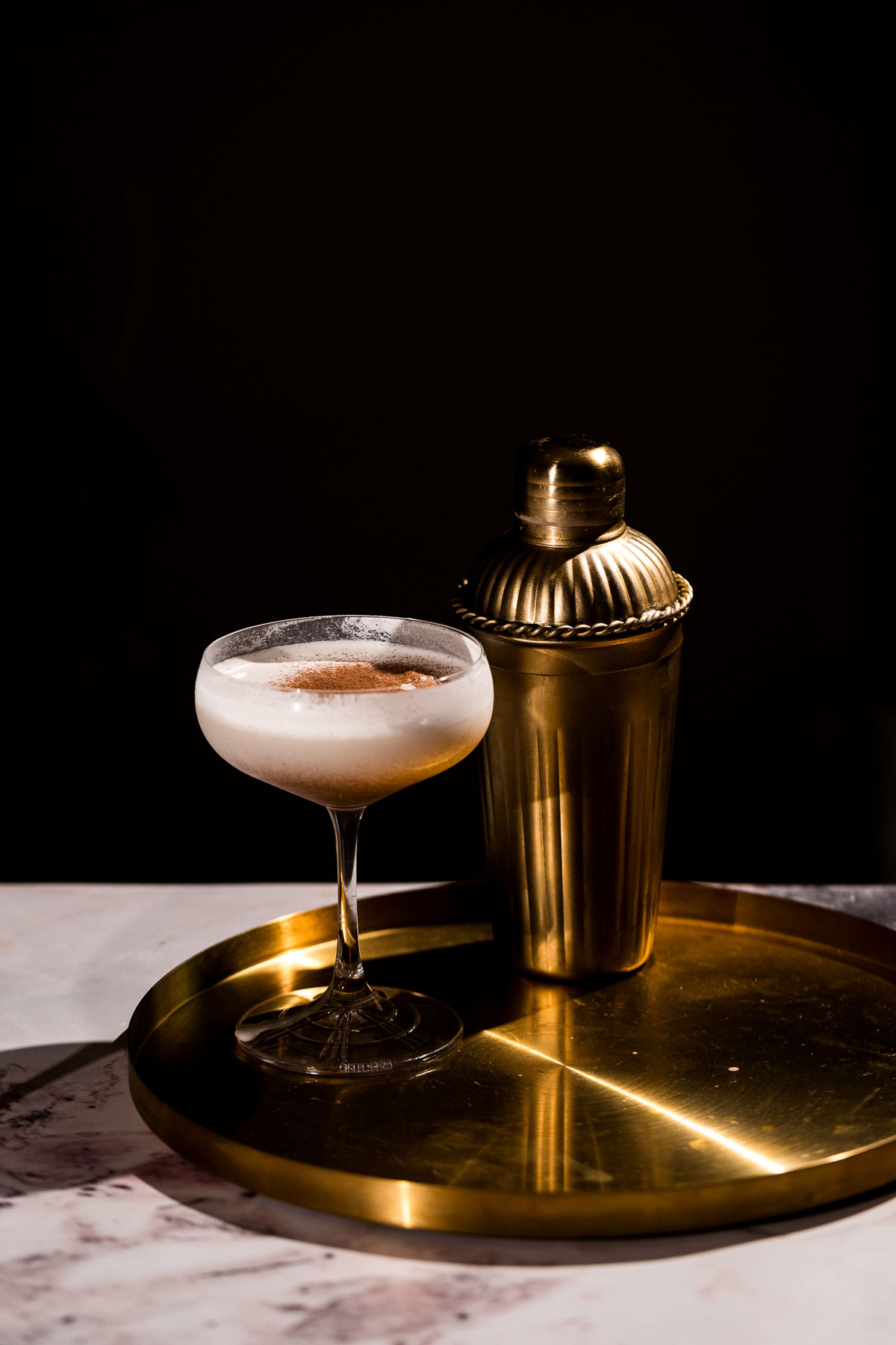 A coupe glass filled with an eggnog martini and a gold shaker rest on a gold tray against a dark background.