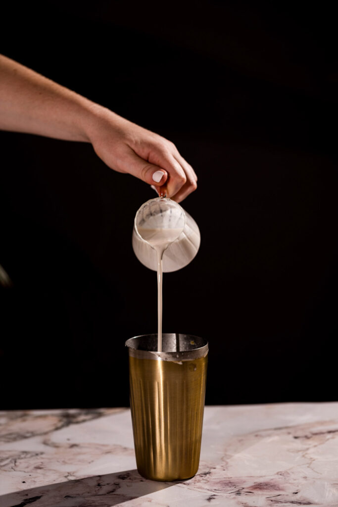 Foamy eggnog is poured from a pitcher into a golden cocktail shaker on marble.