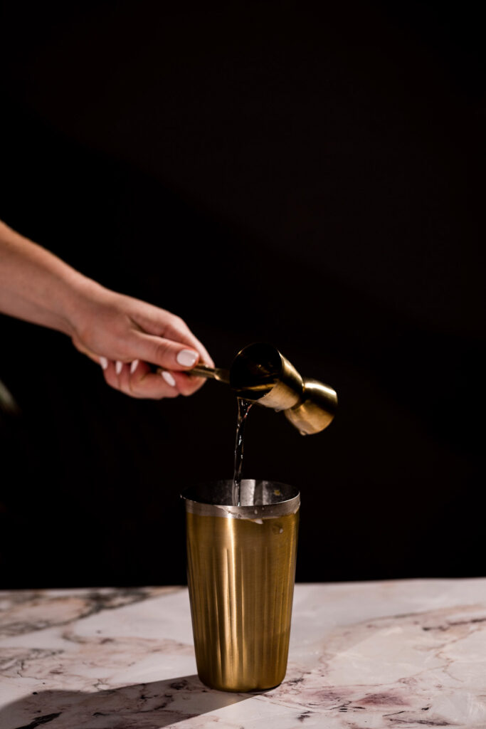 A hand pours vodka from a small brass jigger into a larger brass cup on a marble surface.