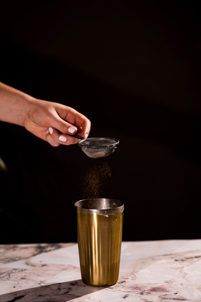 A hand sifts nutmeg over a golden cup on marble.